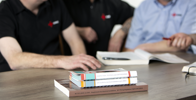 Three contractors are communicating and making notes whilst attending an NICEIC training course. There are training and learning documents stacked up on the contractors table for reference.