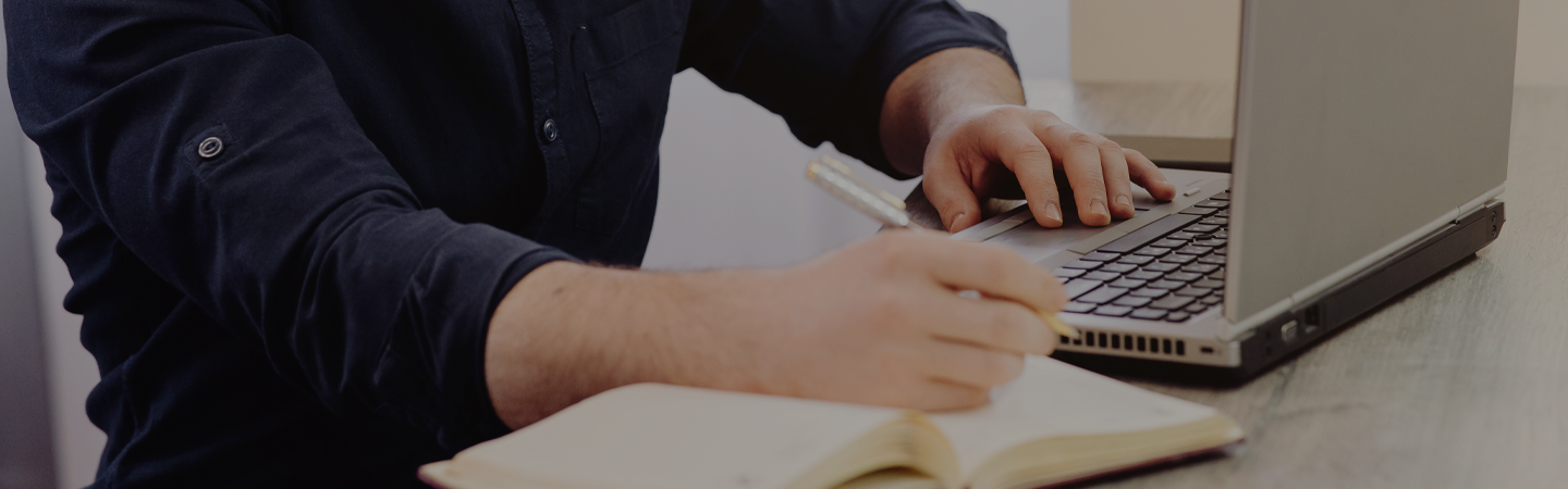 Male working on computer remotely, whilst making notes in a book sat down at a deck.