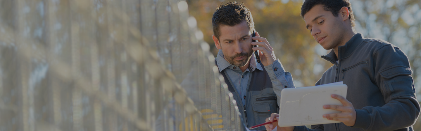 two contractors looking at a digital tablet, whilst one person is on a mobile phone, outside on a construction site.